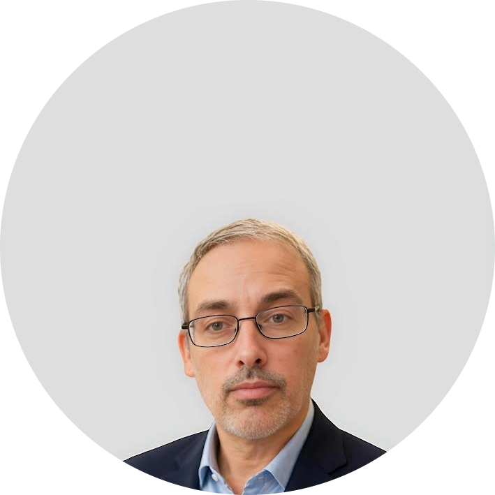 A middle-aged man with short gray hair, glasses, and a trimmed beard, wearing a navy suit and light blue shirt, is centered in front of a plain light gray background.
