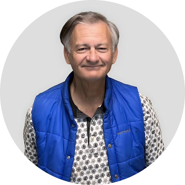 An older man with gray hair smiles at the camera, wearing a bright blue vest over a patterned shirt, standing against a plain light gray background.