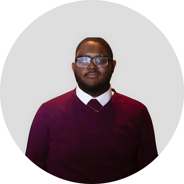 A person wearing glasses, a white collared shirt, and a maroon sweater stands in front of a plain light gray background, facing the camera and smiling slightly.