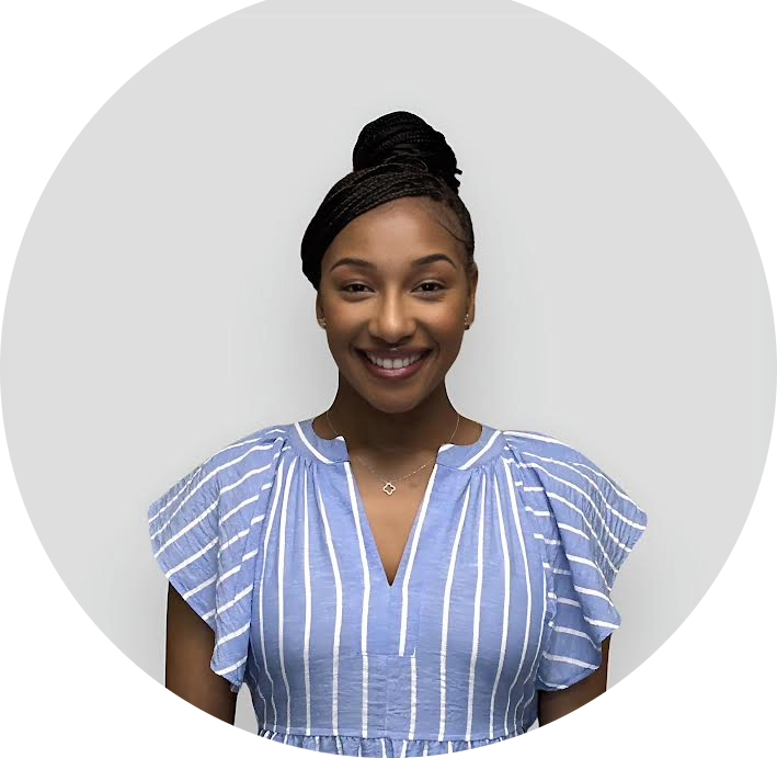 A woman with braided hair in a top bun is smiling. She is wearing a blue and white striped blouse with ruffled sleeves and a delicate necklace. The background is plain and light gray.