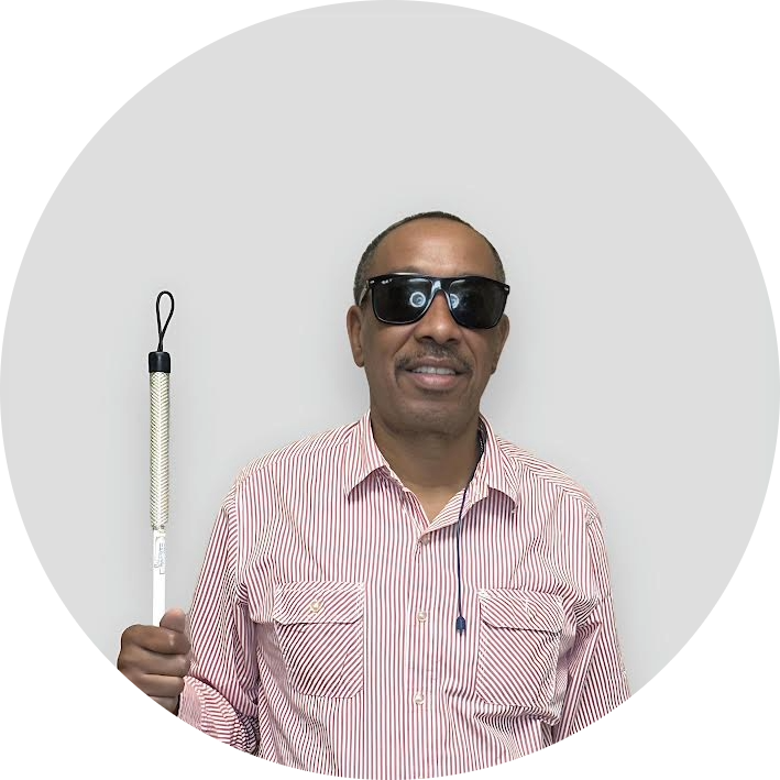 A man wearing dark sunglasses and a striped shirt holds a white cane, standing against a plain light-colored background.
