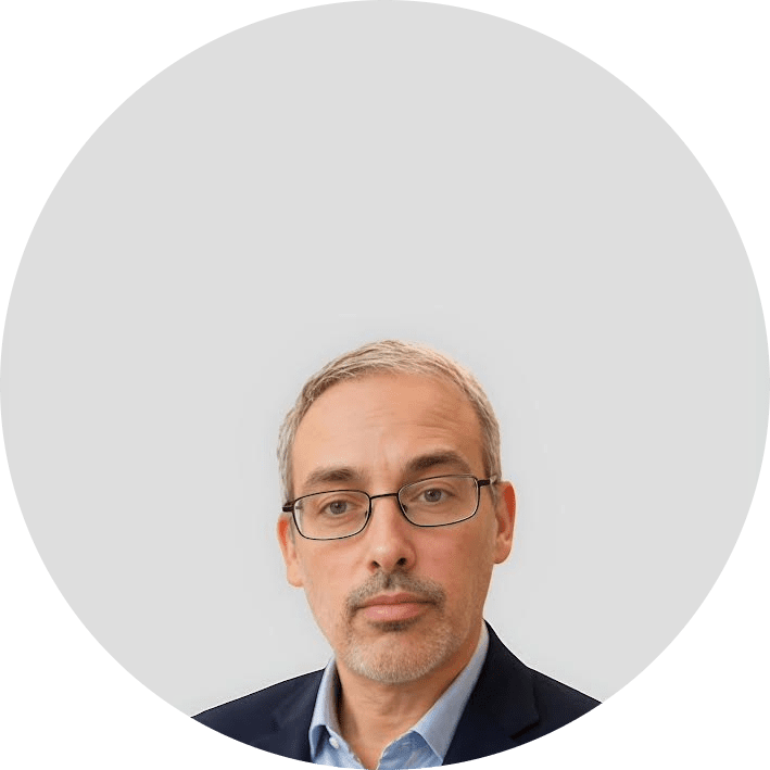 A middle-aged man with short gray hair, glasses, and a trimmed beard, wearing a navy suit and light blue shirt, is centered in front of a plain light gray background.