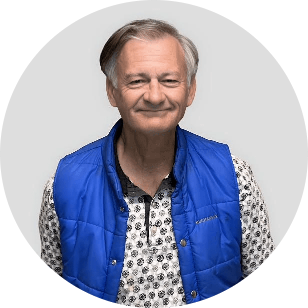 An older man with gray hair smiles at the camera, wearing a bright blue vest over a patterned shirt, standing against a plain light gray background.
