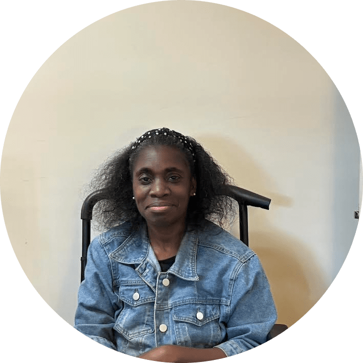 A woman with long curly hair, wearing a blue denim jacket and a black headband, sits and smiles slightly in front of a plain light-colored wall.