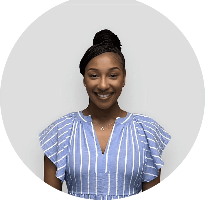 A woman with braided hair in a top bun is smiling. She is wearing a blue and white striped blouse with ruffled sleeves and a delicate necklace. The background is plain and light gray.