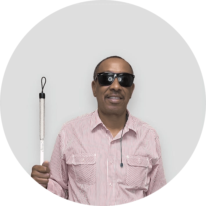 A man wearing dark sunglasses and a striped shirt holds a white cane, standing against a plain light-colored background.