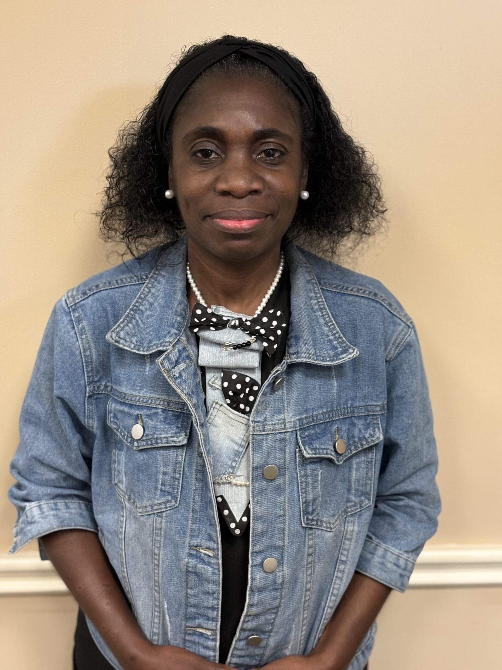 A woman with curly black hair, wearing a denim jacket over a black outfit with a polka dot scarf and pearl necklace, stands in front of a beige wall, smiling gently at the camera.